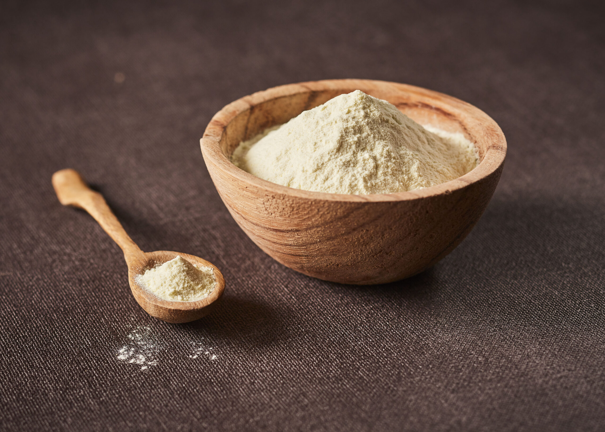 Ramaquin rond en bois contenant la poudre : Milk Salt disposé sur une nappe avec une cuillère en bois à côté contenant aussi cet ingrédient.